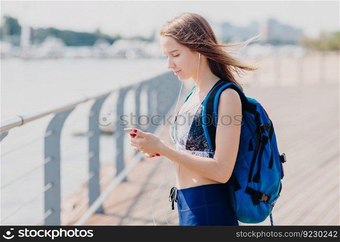 Sideways shot of serious brunette female looks seriously at screen of smart phone, listens electronic book, carries rucksack, poses outdoor, has long walk, breathes fresh air, enjoys calm atmosphere