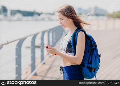 Sideways shot of serious brunette female looks seriously at screen of smart phone, listens electronic book, carries rucksack, poses outdoor, has long walk, breathes fresh air, enjoys calm atmosphere