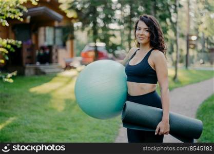 Sideways shot of slim active young woman holds big fitness ball and karemat, going to do exercises outdoor, has morning workout, poses outside against blurred forest background with green grass