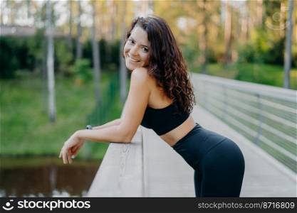 Sideways shot of slim sporty woman wears black top and leggings, has perfect figure, leans at bridge, takes break after doing sport exercising, leads healthy lifestyle. Female runner enjoys nature