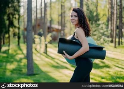 Sideways shot of sporty slim young woman dressed in black leggings, carries rolled up karemat under arm, poses against green grass and trees background, going to have exercises with fitness ball