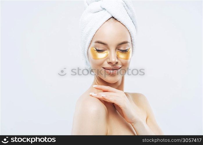 Sideways shot of tender young woman with eye collagen gold pads, healthy fresh skin, anti aging moisturising mask, touches shoulder gently, wears bath towel on head, isolated over white studio wall