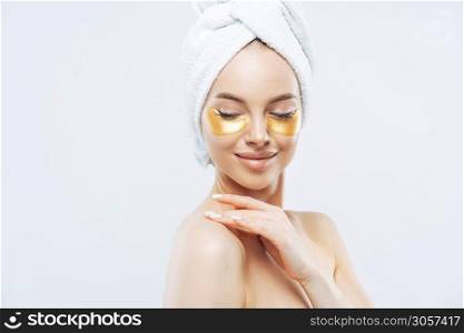 Sideways shot of tender young woman with eye collagen gold pads, healthy fresh skin, anti aging moisturising mask, touches shoulder gently, wears bath towel on head, isolated over white studio wall