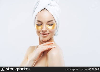 Sideways shot of tender young woman with eye collagen gold pads, healthy fresh skin, anti aging moisturising mask, touches shoulder gently, wears bath towel on head, isolated over white studio wall