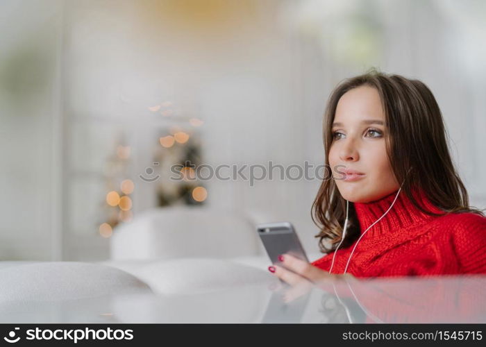 Sideways shot of thoughtful Caucasian woman dressed in knitted sweater, holds modern cell phone in hands, listens audio with earphones, connected to wireless internet, poses in living room alone