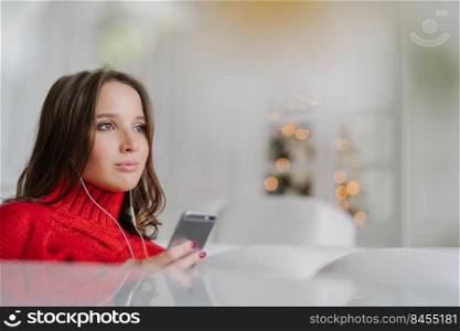 Sideways shot of thoughtful Caucasian woman dressed in knitted sweater, holds modern cell phone in hands, listens audio with earphones, connected to wireless internet, poses in living room alone