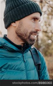 Sideways shot of unshaven man with dark thick beard, wears warm har and anorak, looks pensively aside, poses outside, dreams about something has outdoor stroll in unknown place alone, active lifestyle
