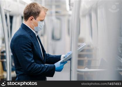 Sideways shot of young elegant man reads newspaper attentively, poses in metro carriage, wears medical mask and disposable rubber gloves from coronavirus. Social distancing, quarantine measures