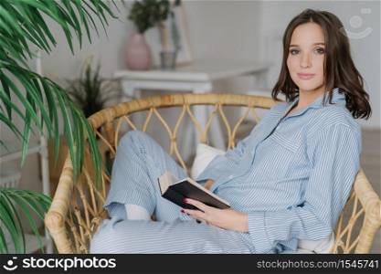 Sideways shot of young female reader hold book in hands, rests at home, wears pyjamas, enjoys calm atmosphere, reading bestseller, sits in wicker chair. People, hobby and relaxation concept.