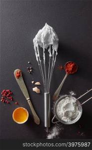 Sieve with flour, knife with red pepper, a metal whisk and halves of a boiled egg on a black concrete background. Concept preparation. Flat lay. A set of knife with pepper, sieve with flour, metal whisk and boiled eggs on a black concrete background. Preparation for cooking. Flat lay