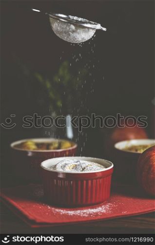 Sifting powdered sugar by sieve over apple pie in ceramic baking molds ramekin on dark wooden table. Close up, shallow depth of the field.