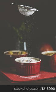 Sifting powdered sugar by sieve over apple pie in ceramic baking molds ramekin on dark wooden table. Close up, shallow depth of the field.