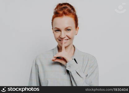 Sign of silence. Headshot portrait of cheerful young ginger 30s woman showing hush gesture, keeping finger on lips while posing isolated on grey studio background. Privacy and silence concept. Cheerful young ginger 30s woman showing hush gesture while posing isolated on grey studio background