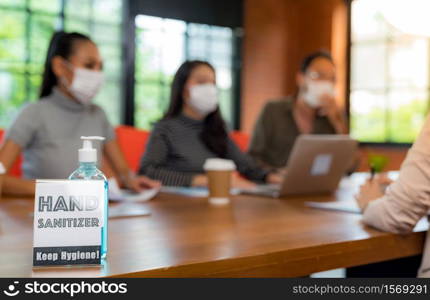 Signage of Hand sanitizer alcohol gel in meeting room with background of Asian team business people working and brainstorm. They wear face mask in new normal office to prevent covid-19 virus spreading