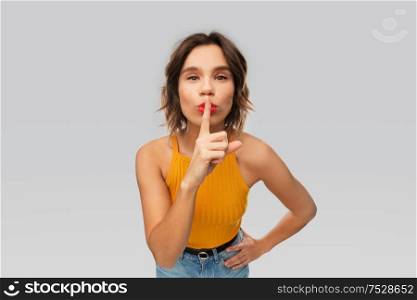 silence, secret, confidentiality concept - young woman in mustard yellow top making hush gesture over grey background. young woman in mustard yellow top