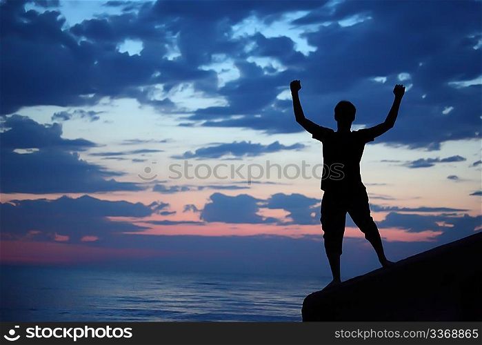 Silhouette guy lifted hands upwards on breakwater in evening near sea