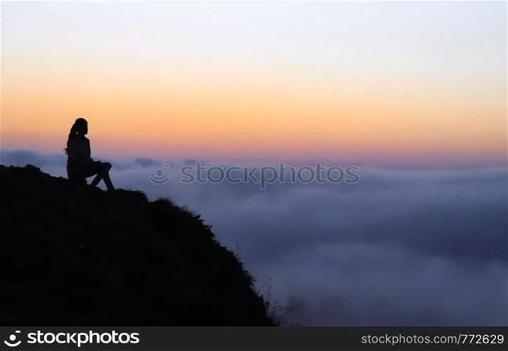 Silhouette landscape of a girl sitting on the edge of a mountain top seeing the clouds float below her as the sunset approaches. A