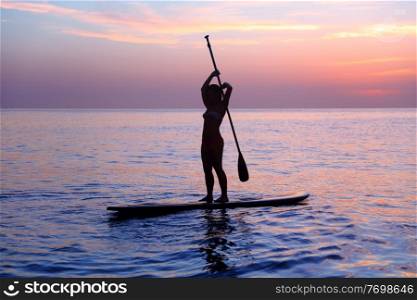 Silhouette of a girl practicing SUP, balancing on paddle board in the sea over beautiful purple sunset, healthy lifestyle and active summer vacation