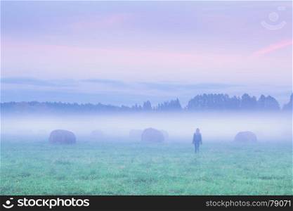 Silhouette of a lonely woman walking away in foggy field with haystacks at sunset