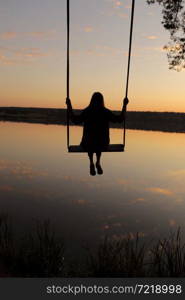 silhouette of a romantic young woman on a swing over lake at sunset. Young girl traveler sitting on the swing in beautiful nature, view on the lake.. silhouette of a romantic young woman on a swing over lake at sunset. Young girl traveler sitting on the swing in beautiful nature, view on the lake