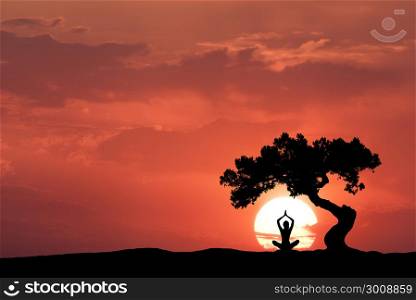 Silhouette of a sitting woman practicing yoga with raised up arms under the crooked tree on the hill on the background of sun and colorful red sky with clouds. Landscape with meditating girl at sunset