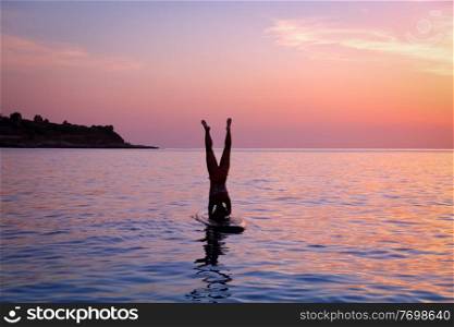 
Silhouette of a sportive woman doing yoga exercise on the sup board over sunset, doing Sirsasana, headstand, active lifestyle, enjoying water sport, active summer vacation