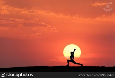 Silhouette of a standing sporty woman practicing yoga. Silhouette of a standing sporty woman practicing yoga with raised up arms on the hill on the background of yellow sun and orange sky with clouds. Landscape with girl at sunset. Fitness and lifestyle