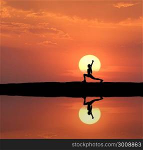 Silhouette of a standing sporty woman practicing yoga with raised up arms on the hill near the lake with sky reflection in water. Sun and orange sky with clouds. Landscape with girl at sunset. Fitness
