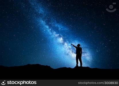 Silhouette of a standing young man pointing finger in night starry sky on the background of blue Milky Way. Colorful night landscape. Beautiful Universe, travel background with sky full of stars