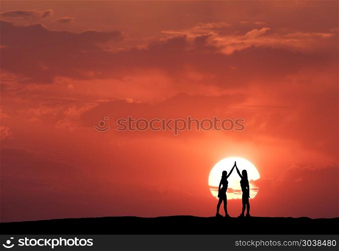 Silhouette of a standing young sporty girls. Silhouette of a standing young sporty girls with raised up arms on the hill on the background of sun and red sky with clouds. Landscape with women holding hands at colorful sunset. Lifestyle