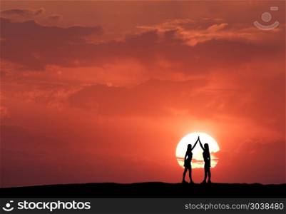 Silhouette of a standing young sporty girls. Silhouette of a standing young sporty girls with raised up arms on the hill on the background of sun and red sky with clouds. Landscape with women holding hands at colorful sunset. Lifestyle
