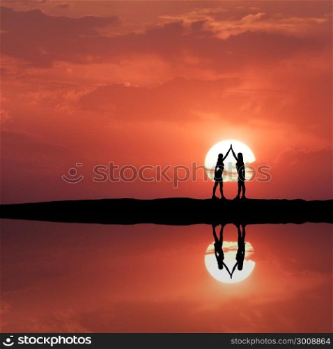 Silhouette of a standing young sporty girls with raised up arms on the hill near the river with sky reflection in water. Sun and red sky with clouds. Landscape with women holding hands at sunset