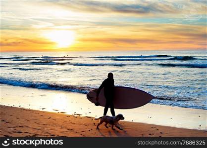 Silhouette of a surfer with a dog walking at sunset on the beach