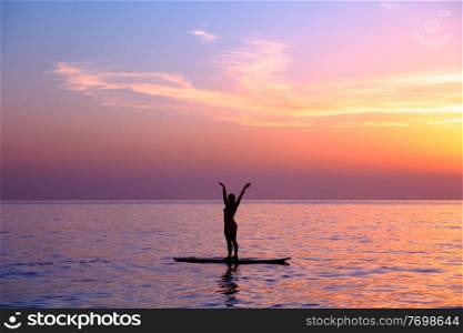 Silhouette of a yoga trainer over sunset background doing asanas, balancing on the sup board, enjoying healthy lifestyle, summer vacation on the beach