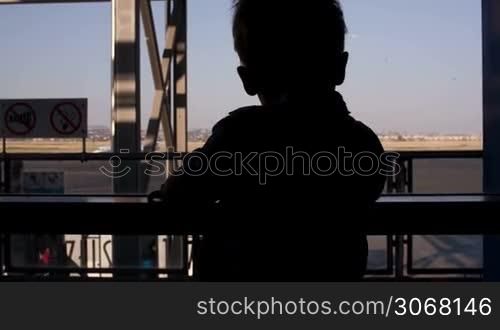 Silhouette of a young boy looking through a window in the airport to the passengers passing by and airplanes