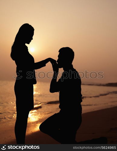 Silhouette of boyfriend kneeling and kissing his girlfriends hand on the beach at sunset