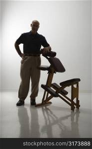 Silhouette of Caucasian middle-aged male massage therapist standing with elbow on massage chair.