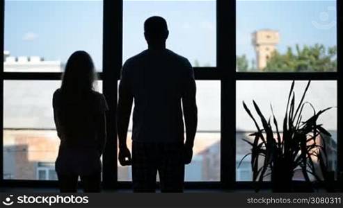 Silhouette of cheerful family with little daughter embracing against big window at home. Joyful father taking cute girl in his arms and hugging his attractive wife while family standing in front of the window. Back view.