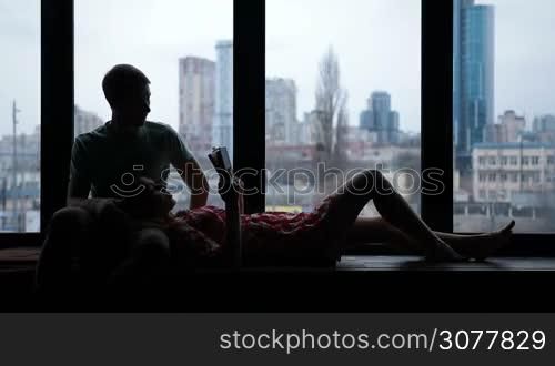 Silhouette of couple in love spending leisure together and reading book on windowsill at home over cityscape background. Beautiful woman lying on back, with head on man&acute;s lap, reading a book on windowsill. Happy family lounging together in room.
