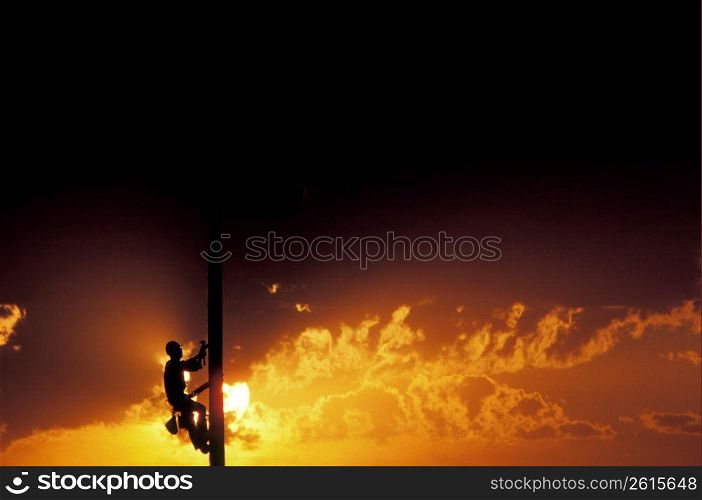 Silhouette of electrician climbing telephone pole with sunset in background