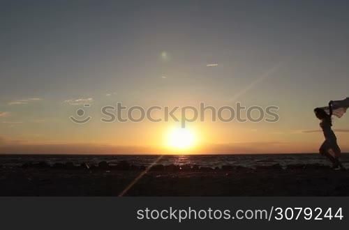 Silhouette of happy woman running with flying scarf on the beach against dramatic sky during sunset. Carefree long hair female in summer dress with flying scarf in hands jogging on the sea shore in glow of orange setting sun. Slow motion.