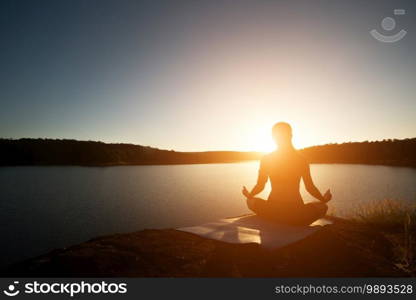 Silhouette of healthy woman is practicing yoga at mountain lake during sunset. 