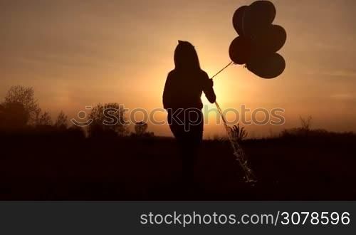 Silhouette of little girl with colorful balloons walking in the meadow in glowing of sunset. Rear view. Cute child with many big balloons stepping ahead in the rays of sunset in the field. Slow motion. Steadicam stabilized shot.
