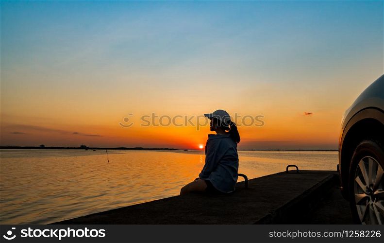 Silhouette of lonely young woman wear a cap relaxing on the beach alone in front of the car with orange and blue sky at sunset. Summer vacation and travel concept.