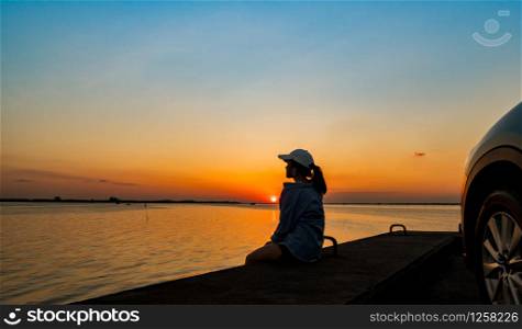 Silhouette of lonely young woman wear a cap relaxing on the beach alone in front of the car with orange and blue sky at sunset. Summer vacation and travel concept.
