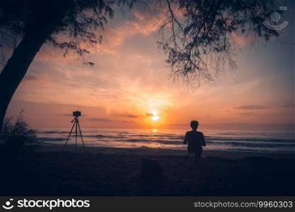 silhouette of man enjoying the sunrise at the beach and using smartphone on tripod to take pictures.