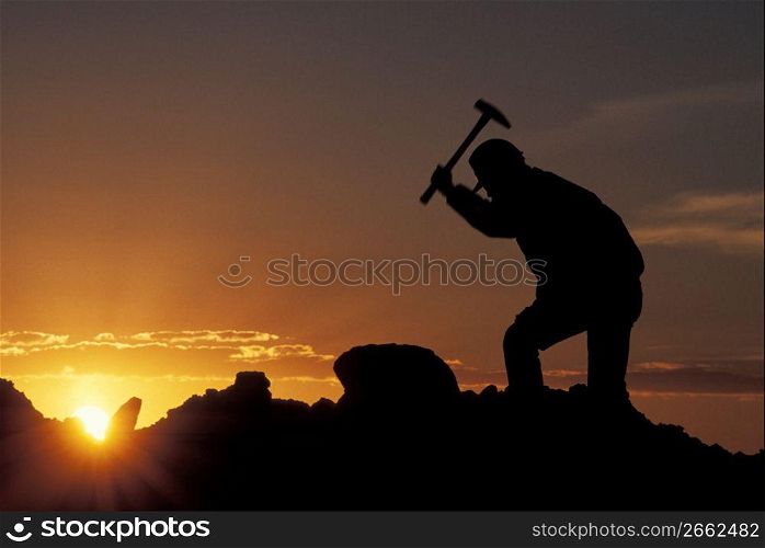 Silhouette of man hitting pile of rocks with hammer while yellow sun sets in the background