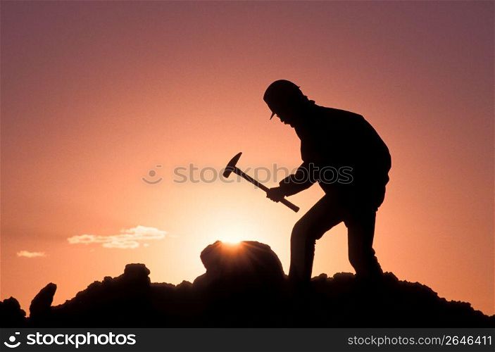 Silhouette of man hitting pile of rocks with hammer while yellow sunset in the background