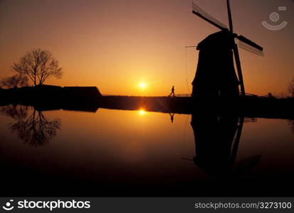 Silhouette of man running and old-fashioned, wooden windmills along canal with yellow setting sun in background