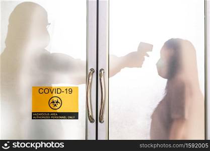 Silhouette of medical staff with PPE suit taking temperature to asian woman before do coronavirus test with signage of COVID-19 quarantine zone. New normal healthcare service and medical concept.
